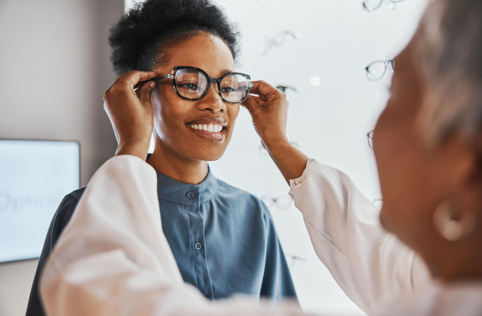 A young woman smiling and trying on glasses in a store while being assisted by an optometrist.