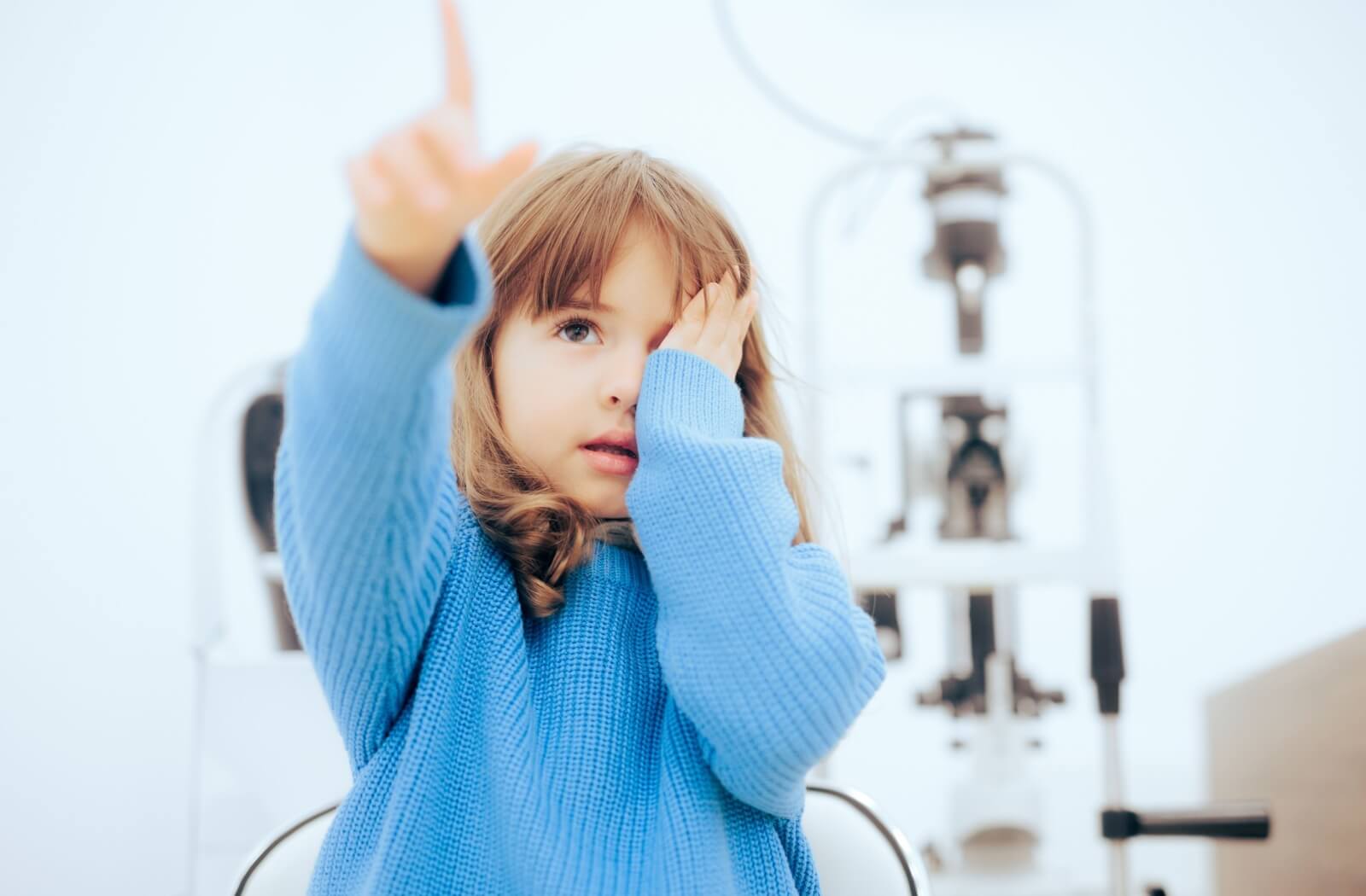 A little girl in a blue sweater covering one eye and pointing during an eye exam.