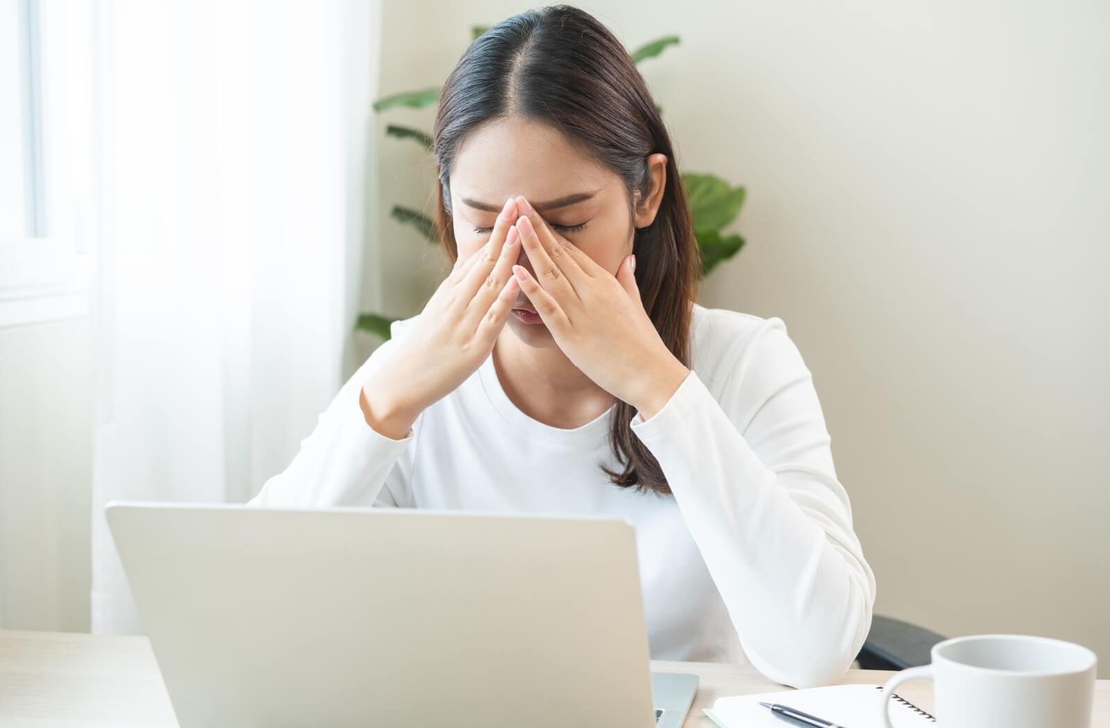 A person working at their laptop pinches their eyes due to discomfort from dry eye.