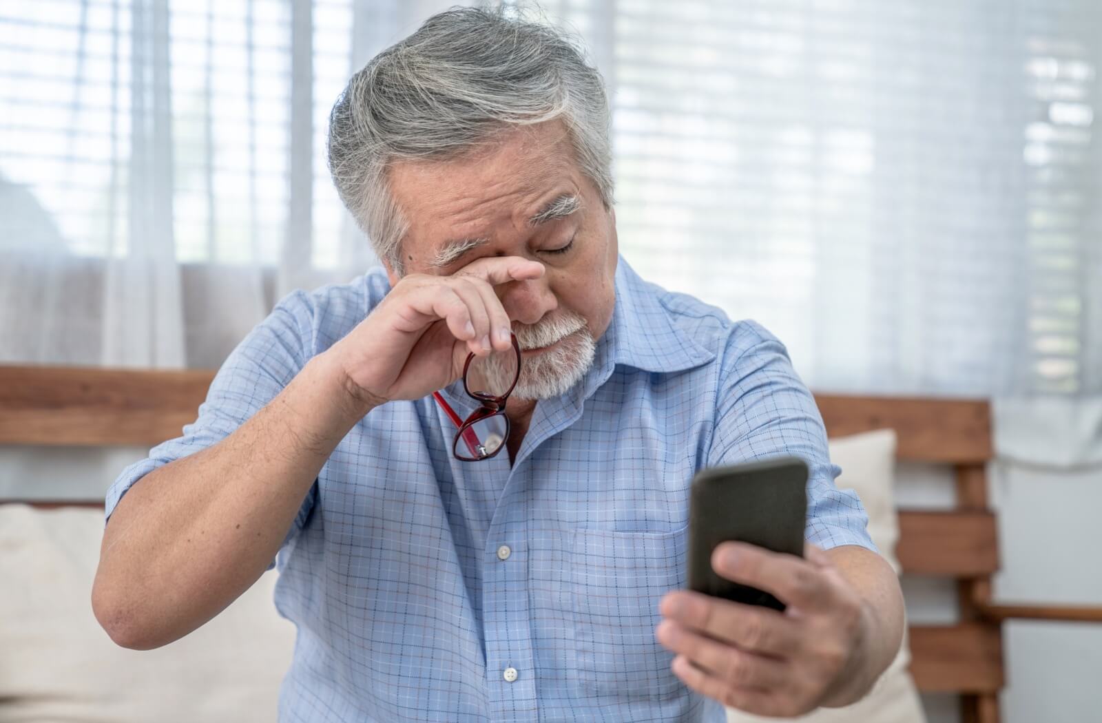 A senior sits on a bed in a well-lit room, rubbing their eyes with their right hand while holding their phone at arm's length, struggling with vision issues from glaucoma.