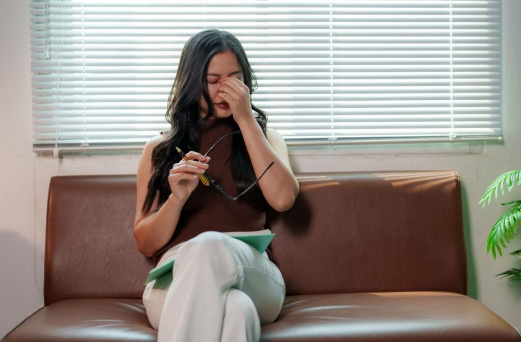 Person sitting on couch rubbing eyes while holding glasses showing discomfort from dry eyes
