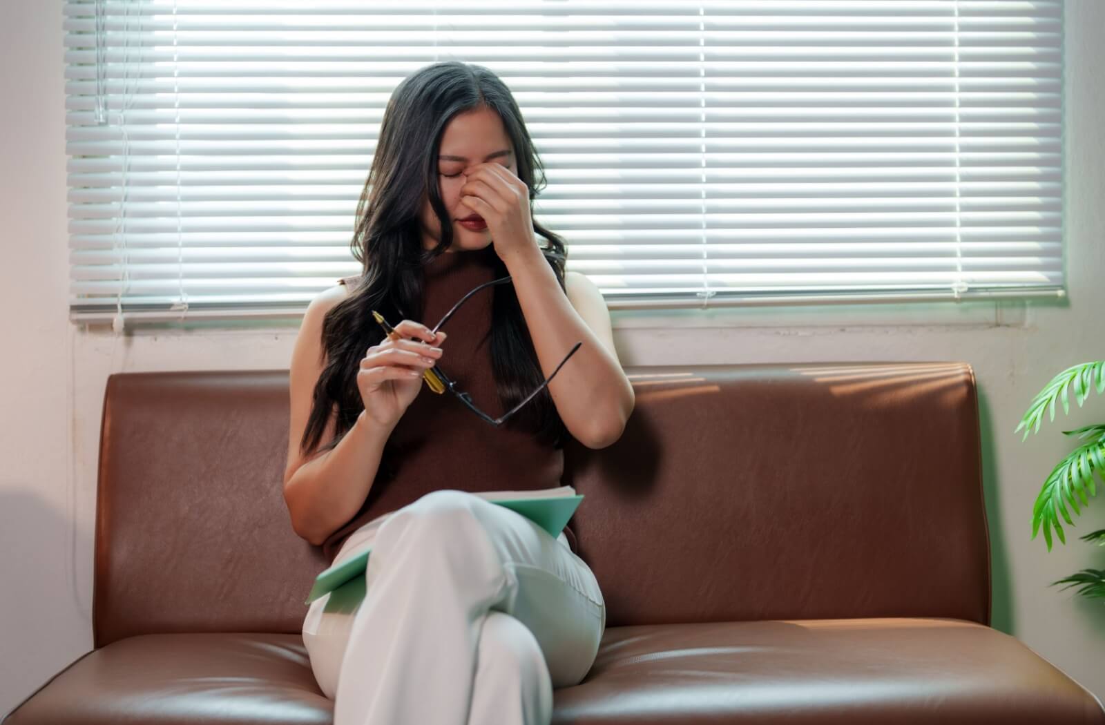 Person sitting on couch rubbing eyes while holding glasses showing discomfort from dry eyes