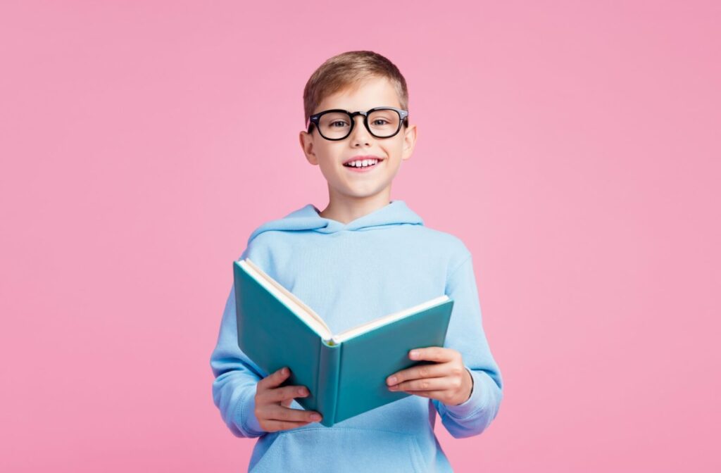 Boy wearing glasses and holding a book against a pink background.