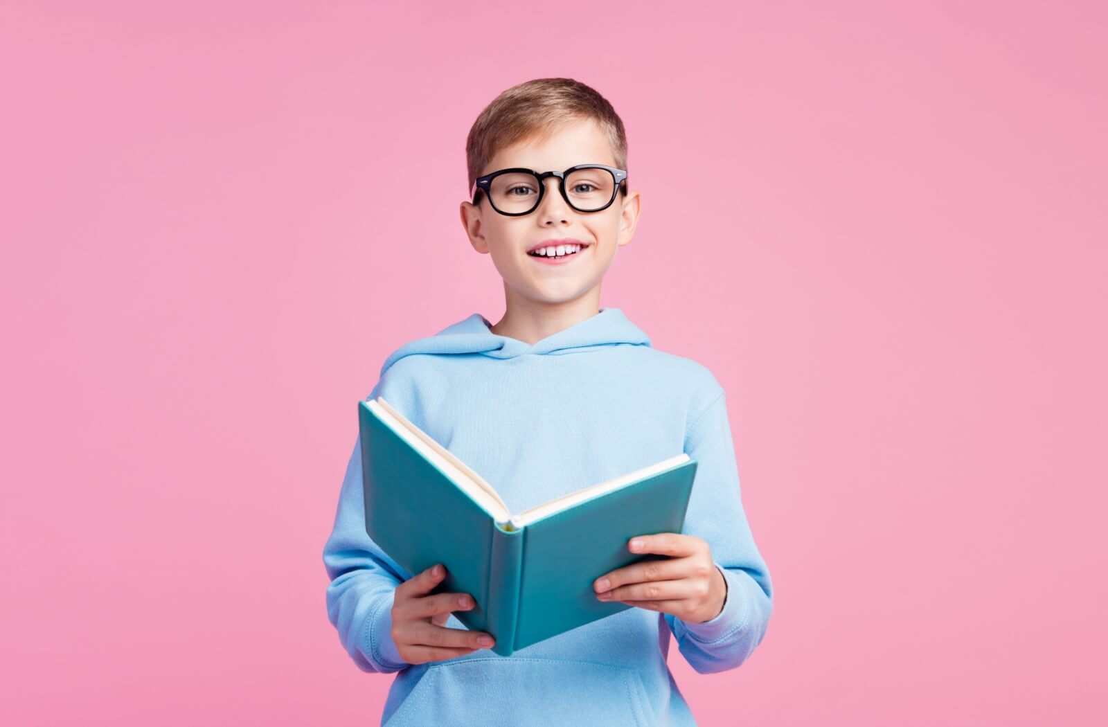 Boy wearing glasses and holding a book against a pink background.