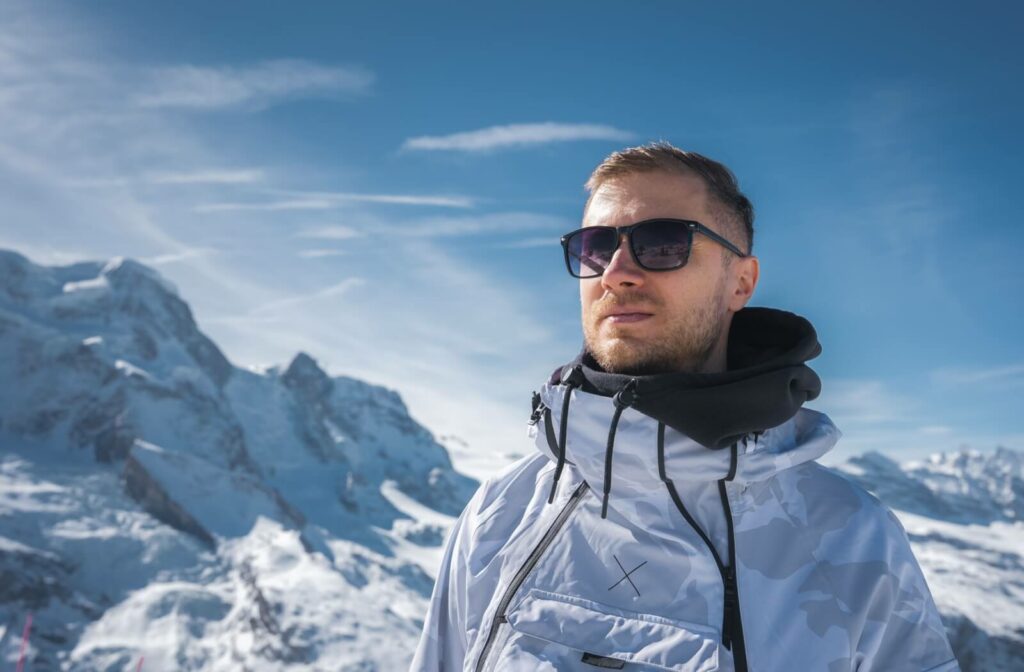 Person wearing sunglasses and a winter jacket standing outdoors with snowy mountains and a clear blue sky in the background.