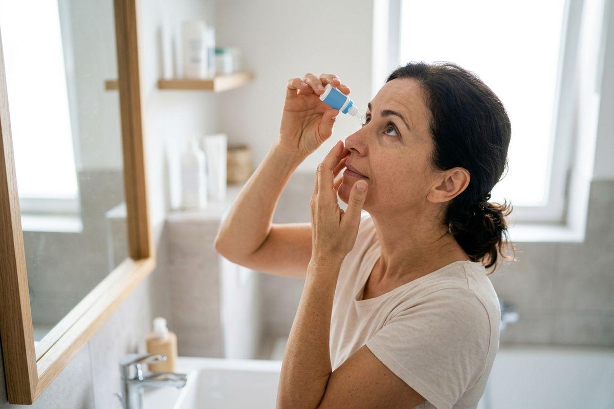 An adult in a bathroom looks up, pulling down their lower eyelid to squeeze drops from a small blue bottle into their eye.